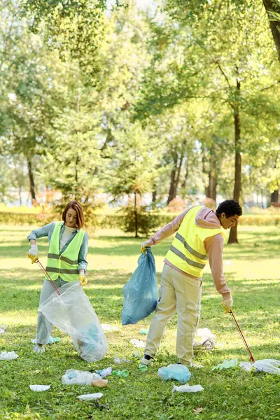 A diverse couple in safety vests and gloves standing in the grass, cleaning a park together with love and dedication. — Stock Photo