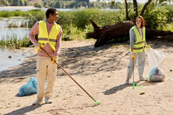 A diverse couple, wearing safety vests and gloves, stand in the sand together, cleaning the park. — Stock Photo