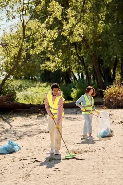 Una coppia eterogenea socialmente attiva in giubbotti e guanti di sicurezza si trova nella sabbia, incarnando amore e lavoro di squadra mentre puliscono il parco.. — Foto stock