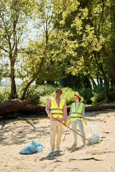 A socially active, diverse, and loving couple in safety vests and gloves cleaning a park together while standing in the sand. — Stock Photo