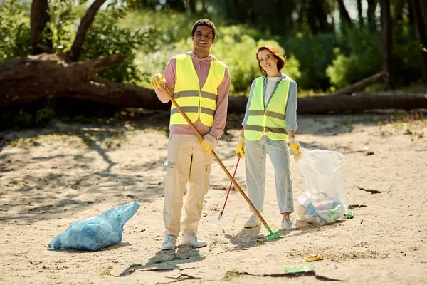 A diverse couple in safety vests and gloves stand on the sandy beach, united in cleaning the environment, showing love and care. — Stock Photo