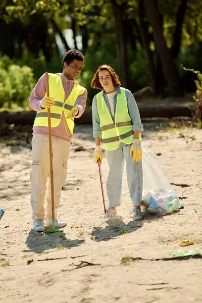Una pareja socialmente activa y diversa que usa chalecos y guantes de seguridad, de pie en la arena mientras limpia el parque. - foto de stock