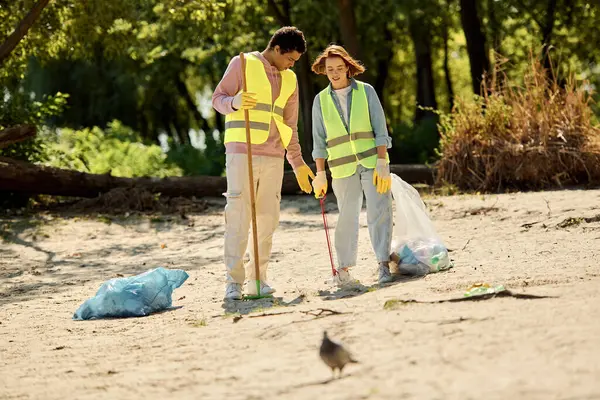 Una coppia eterogenea, indossando giubbotti di sicurezza e guanti, pulisce un parco mentre si trovano insieme nella sabbia. — Foto stock