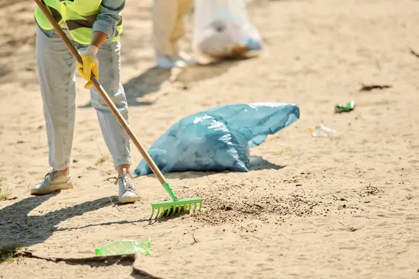 Жінка в жилеті і рукавичках піднімає сміття на пляжі, втілюючи дух екологічного управління і догляду. — стокове фото