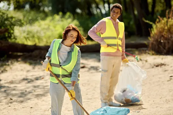 Una coppia eterogenea che indossa giubbotti di sicurezza e guanti si trova nello sporco mentre pulisce un parco insieme. — Foto stock