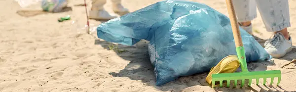 Una pala y una bolsa azul descansan en una playa de arena, simbolizando los esfuerzos ambientales de una pareja socialmente activa que limpia la costa. - foto de stock