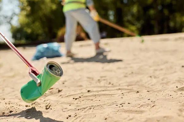 Una lata de refresco se sienta en la playa de arena, con una figura solitaria en el fondo limpiando activamente. - foto de stock
