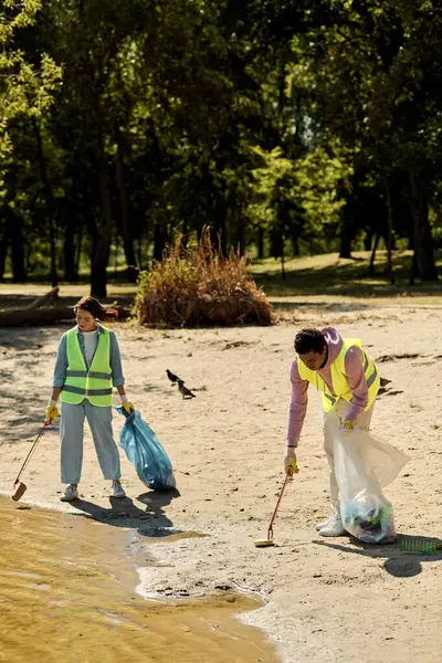 Una pareja socialmente activa y diversa en chalecos y guantes de seguridad, de pie en la arena, limpiando el parque juntos. - foto de stock