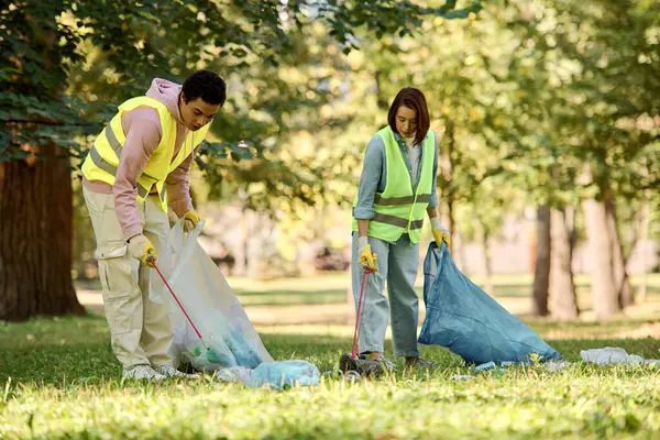 Un couple diversifié et aimant, vêtu de gilets de sécurité et de gants, debout dans l'herbe verte pendant qu'ils nettoient le parc ensemble. — Photo de stock
