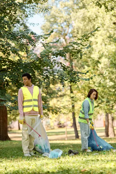 Социально активная, любящая пара в защитных жилетах и перчатках, стоящая в траве, убирающая парк вместе. — стоковое фото