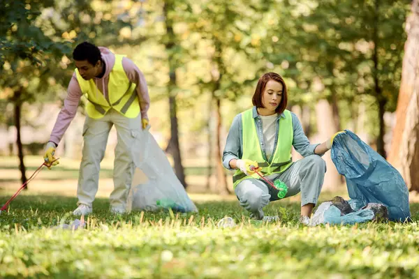 Diverse couple in safety vests and gloves stand on grassy field, actively participating in a park clean-up event. — Stock Photo