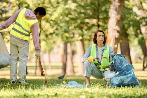 Un couple aimant et diversifié dans des gilets de sécurité et des gants nettoyant un parc ensemble, debout dans l'herbe entourée par la nature. — Photo de stock