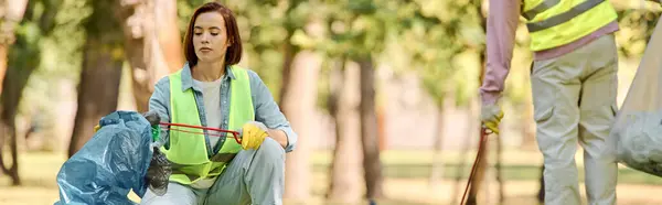 A woman, part of a socially active diverse loving couple, in a safety vest is picking up trash in a park. — Stock Photo
