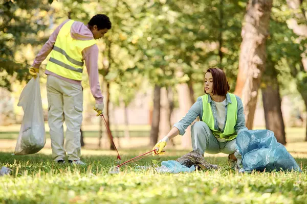 Una pareja socialmente activa y diversa en chalecos de seguridad y guantes limpiando el césped en un parque. - foto de stock