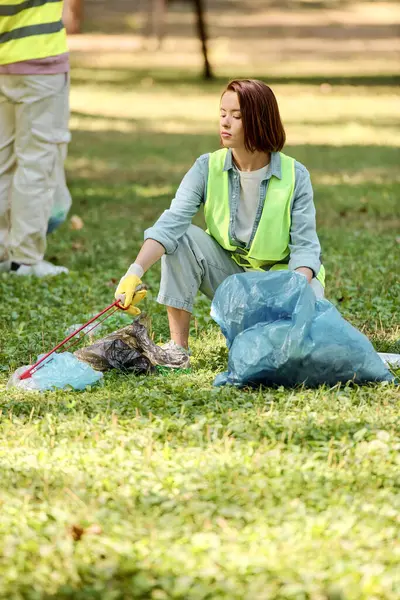 Женщина в защитном жилете и перчатках сидит в траве, держа мешок мусора, очищая окружающую среду с преданностью. — стоковое фото