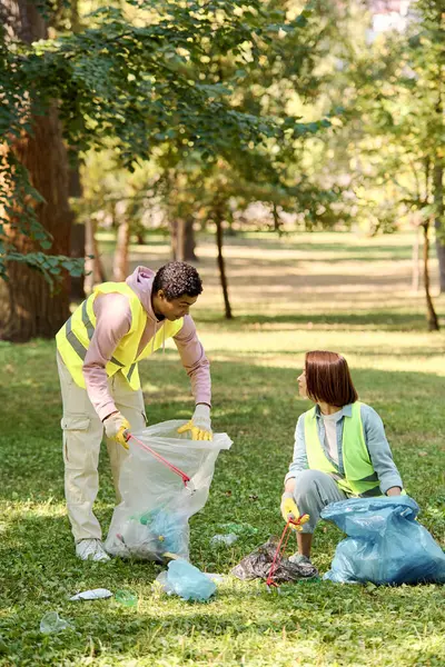Hombre afroamericano y mujer caucásica en chalecos y guantes de seguridad trabajan juntos para recolectar basura en un parque, promoviendo la ecología y el cuidado comunitario. - foto de stock