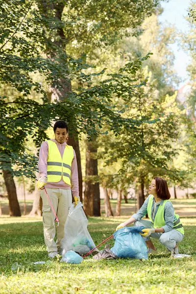 A diverse couple in safety vests and gloves standing in the grass, cleaning a park together. — Stock Photo