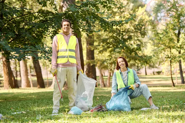 Разнообразная пара в защитных жилетах и перчатках очищает парк, стоящий в траве, объединенный в своей преданности окружающей среде. — стоковое фото