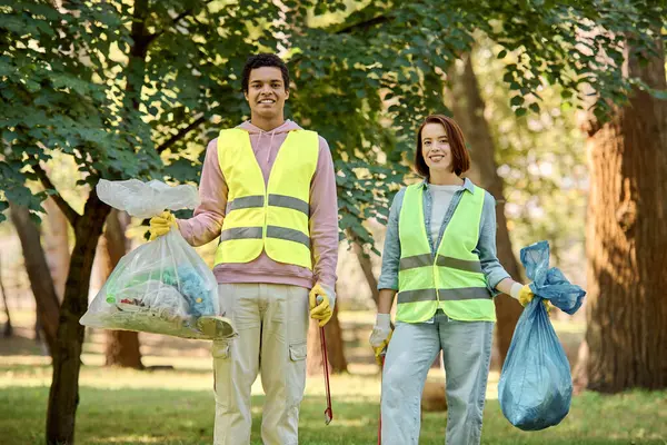 A socially active diverse loving couple in safety vests and gloves, holding bags of garbage while cleaning up the park together. — Stock Photo