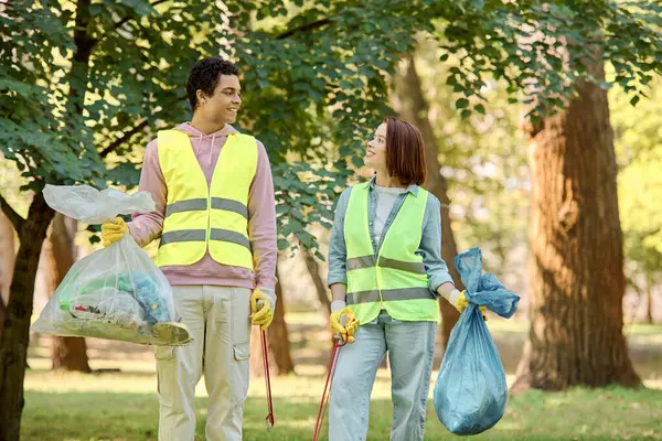 Una pareja socialmente activa y diversa en chalecos de seguridad y guantes limpiando un parque juntos, de pie en la exuberante hierba verde. - foto de stock