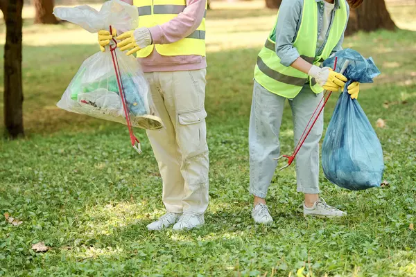 A socially active, diverse couple in safety vests and gloves cleaning a park together, standing in the lush green grass. — Stock Photo