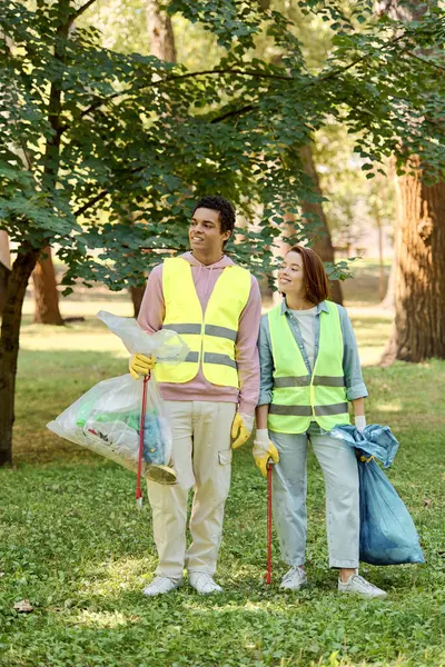 A socially active, diverse couple in safety vests and gloves stand in the grass, cleaning the park together in harmony. — Stock Photo