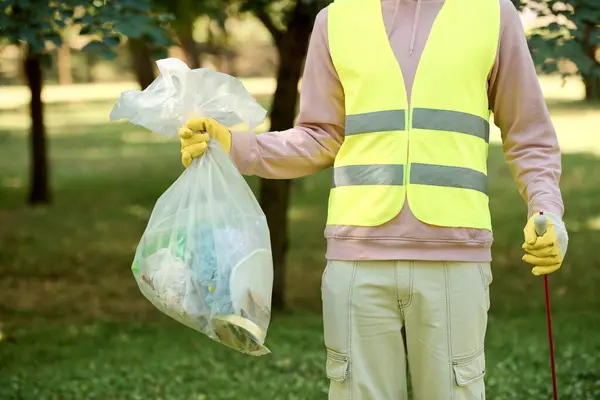 Hombre afroamericano en un chaleco de seguridad de color amarillo brillante sostiene una bolsa de basura mientras participa en una actividad de limpieza del parque. - foto de stock