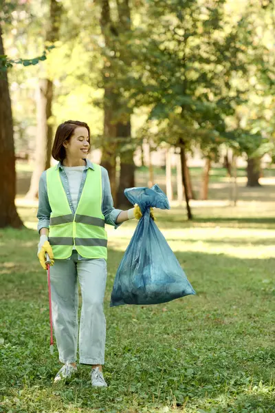 Eine Frau mit grüner Weste und blauem Sack steht selbstbewusst in einem Park, möglicherweise bereit, Müll zu beseitigen. — Stockfoto