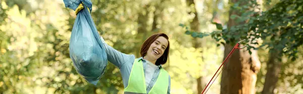 A woman in a green vest holding a blue bag, participating in a park cleaning activity with her partner. — Stock Photo