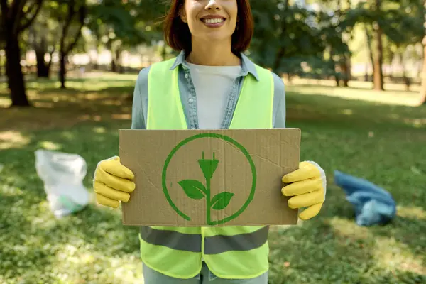A woman in a safety vest carefully holds a cardboard box with a green plant on it while engaging in sustainable gardening. — Stock Photo
