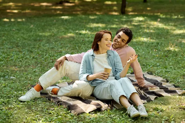 Una coppia eterogenea vestita con abiti vivaci si siede su una coperta nell'erba, godendo di un momento di pace insieme nel parco.. — Foto stock
