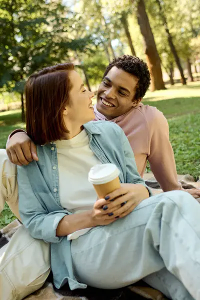 Un uomo e una donna, vestiti con abiti vibranti, si siedono su una coperta con in mano tazze di caffè in un parco. — Foto stock