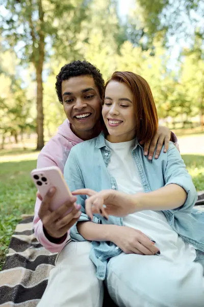Una pareja en traje vibrante sentada en una manta en un parque, absorta en un teléfono celular. - foto de stock