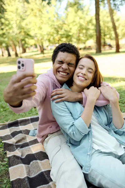 Un homme en tenue vibrante prenant un selfie avec une femme sur une couverture dans le parc, profitant d'un moment d'amour ensemble. — Photo de stock