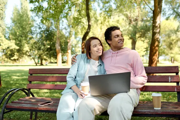 Um homem e uma mulher, vestidos com trajes vibrantes, sentam-se em um banco de parque com um laptop, envolvidos em atividades on-line juntos. — Fotografia de Stock