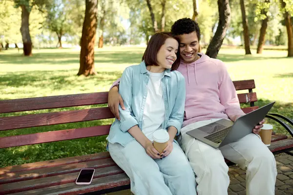 Un couple diversifié en tenue vibrante assis sur un banc de parc, absorbé dans un écran d'ordinateur portable, profitant d'un moment de connexion et de technologie. — Photo de stock