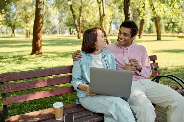 A diverse couple in vibrant attire sits on a park bench, using a laptop together, immersed in their digital world while surrounded by nature. — Stock Photo