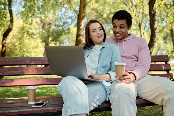 Un couple élégant, vêtu d'une tenue vibrante, s'assoit étroitement sur un banc, absorbé par un travail collaboratif sur un ordinateur portable dans un parc. — Photo de stock