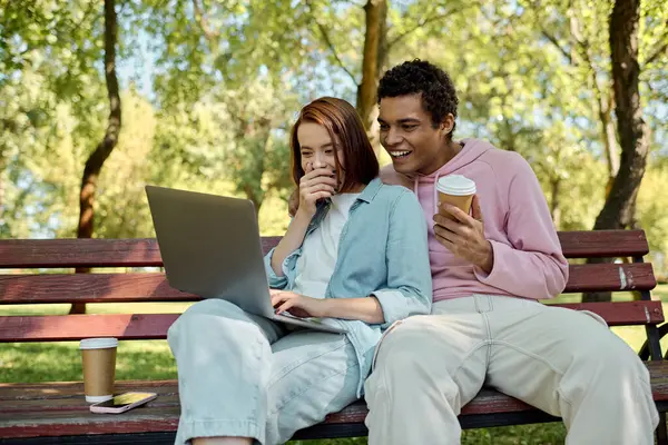 A couple, dressed in vibrant attire, relaxes on a bench with a laptop in a peaceful park setting. — Stock Photo