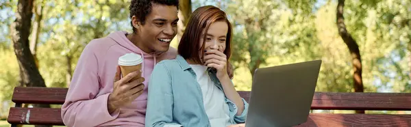 Couple diversifié en tenue vibrante assis sur le banc du parc, absorbé dans l'écran d'ordinateur portable. — Photo de stock