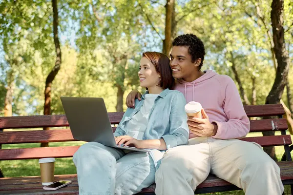A stylish man and woman in colorful outfits sit on a bench, using a laptop outdoors in a park. — Stock Photo