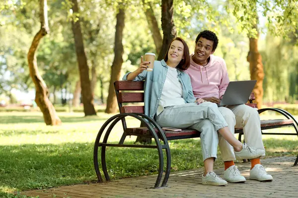 Різноманітна пара сидить на лавці парку, працюючи разом на ноутбуці, оточеному природою. — стокове фото