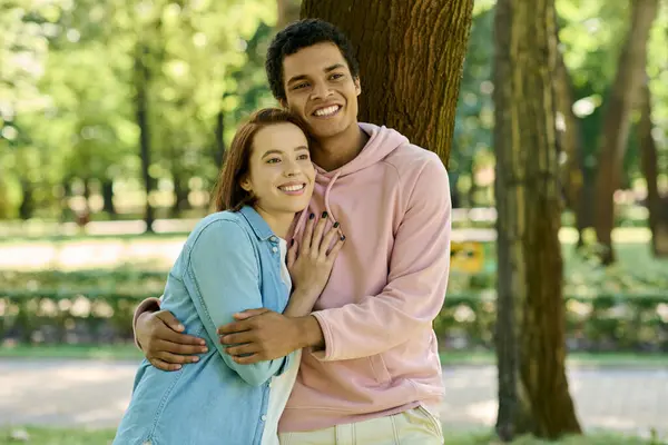 Un hombre y una mujer, una pareja amorosa, vestidos con vibrantes atuendos, de pie juntos junto a un majestuoso roble en un parque. - foto de stock
