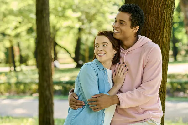 A loving, diverse couple in vibrant attire stand together next to a tree in a park. — Stock Photo