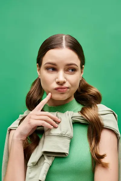 A stylish young woman in her 20s pointing at cheek in a studio setting, wearing a striking green sweater. — Stock Photo