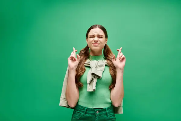 A young woman in her 20s holding fingers crossed in front of her face in a studio setting with a green backdrop. — Stock Photo