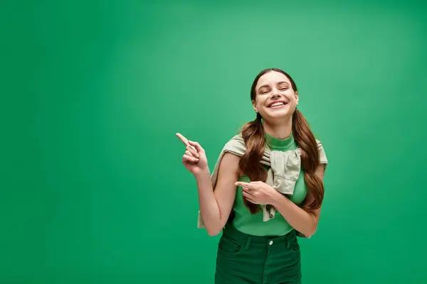 A young woman in her 20s smiles brightly as she points excitedly at something off-camera in a studio setting with a green background. — Stock Photo