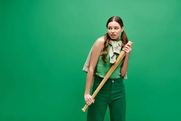 A young beautiful woman in her 20s confidently holds a baseball bat in front of a vibrant green background. — Stock Photo