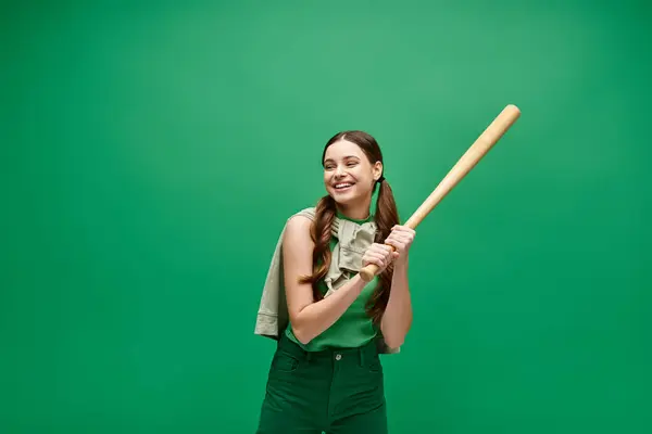 A young woman in her 20s holds a baseball bat confidently against a vibrant green background, exuding strength and determination. — Stock Photo