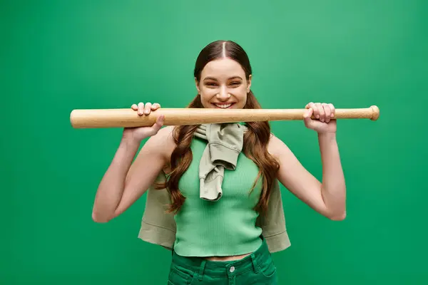 Una giovane, bella donna sulla ventina tiene una mazza da baseball sopra la testa in una posa dinamica sullo sfondo di uno studio verde. — Foto stock